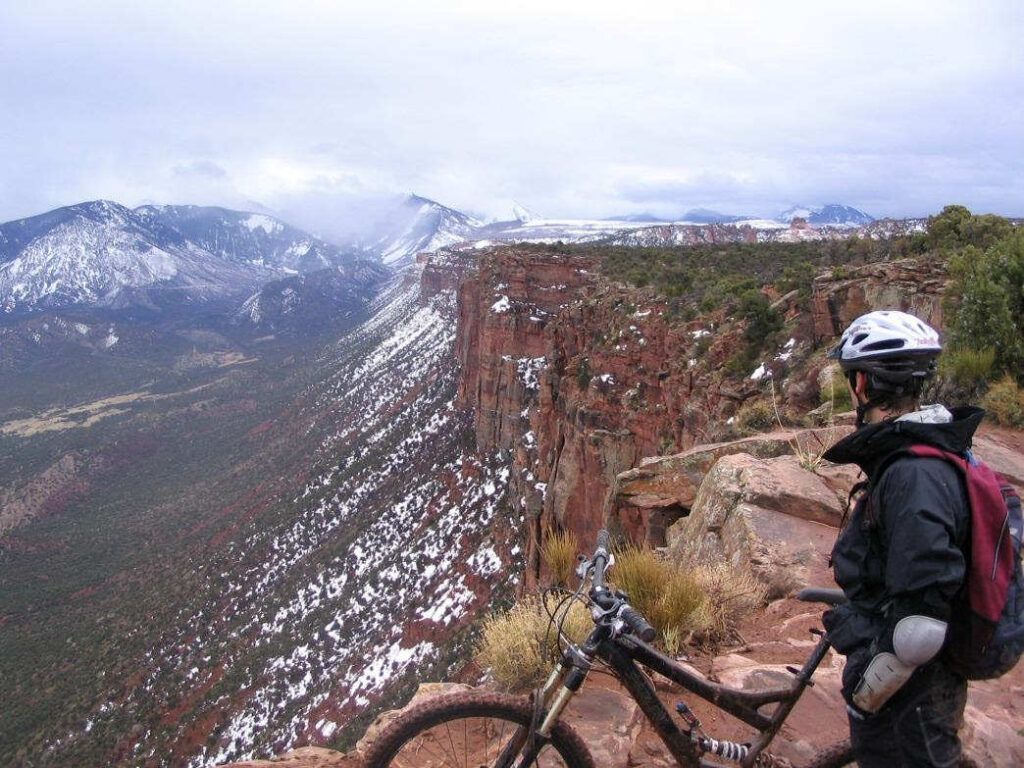 Cyclist with mountain bike on a rocky cliff overlooking a snowy mountain valley