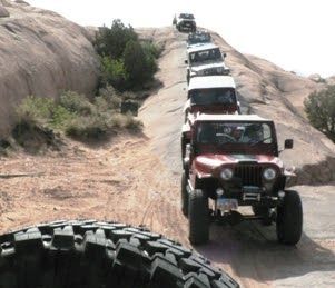 Line of off-road Jeeps climbing a rocky trail in the desert, with a large tire in the foreground