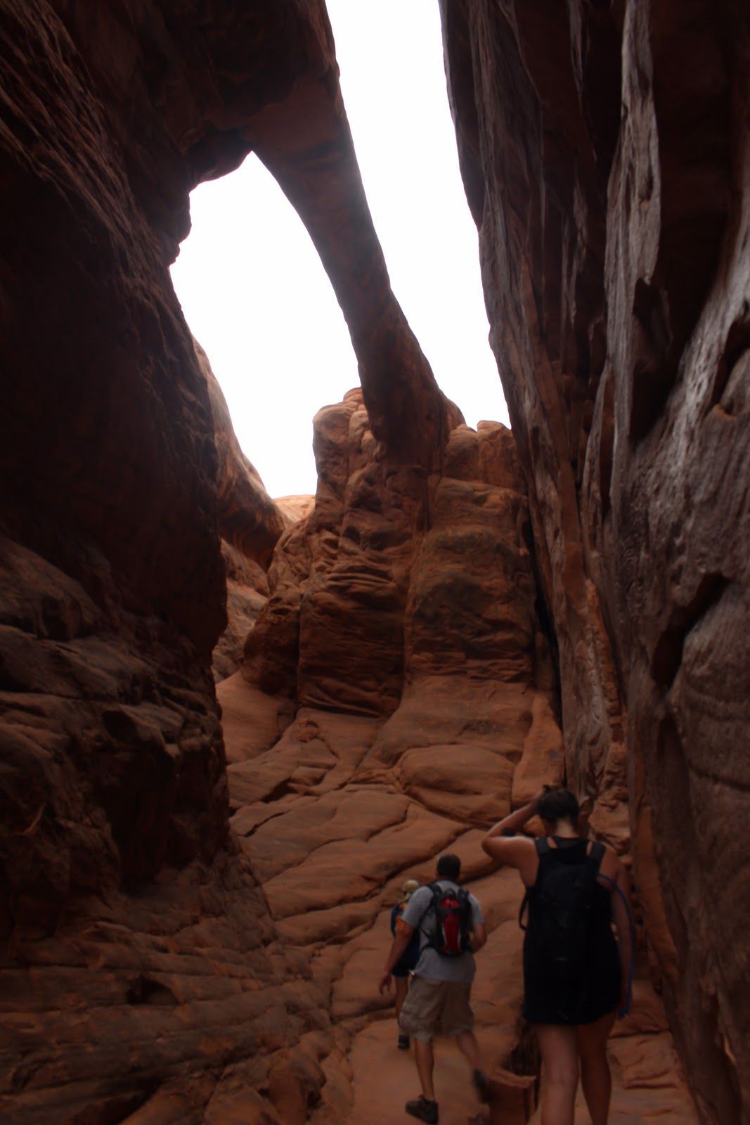 Two hikers climb a narrow red sandstone slot canyon with steep rock walls overhead.