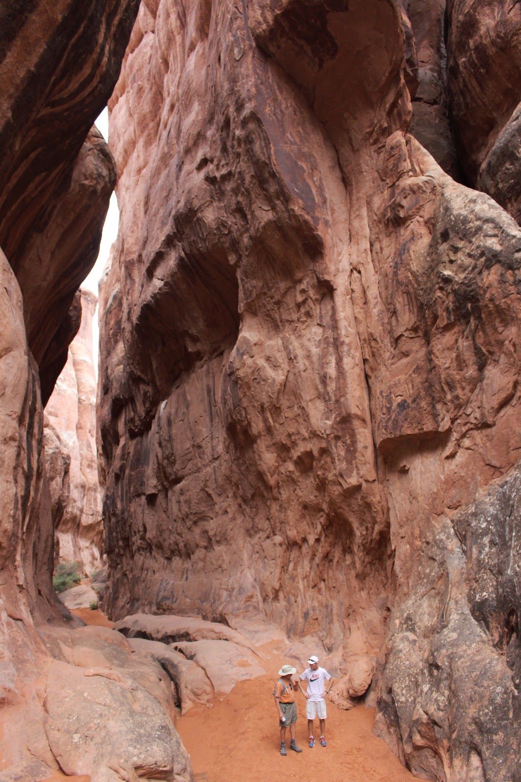Two hikers standing beneath towering red sandstone cliffs in a narrow canyon.