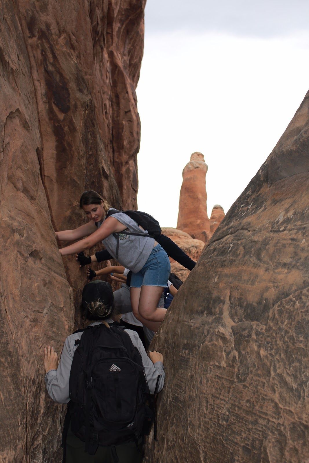 Hikers scrambling through a narrow sandstone slot canyon with a tall rock spire in the background