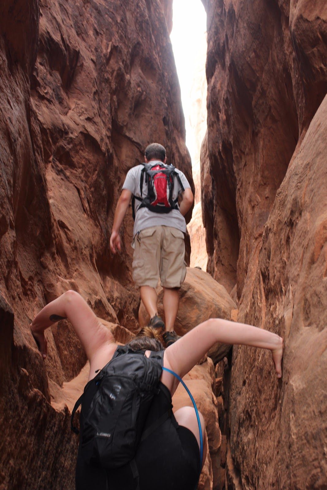 Two hikers in a narrow red-rock canyon, one climbing ahead and one below with a backpack.