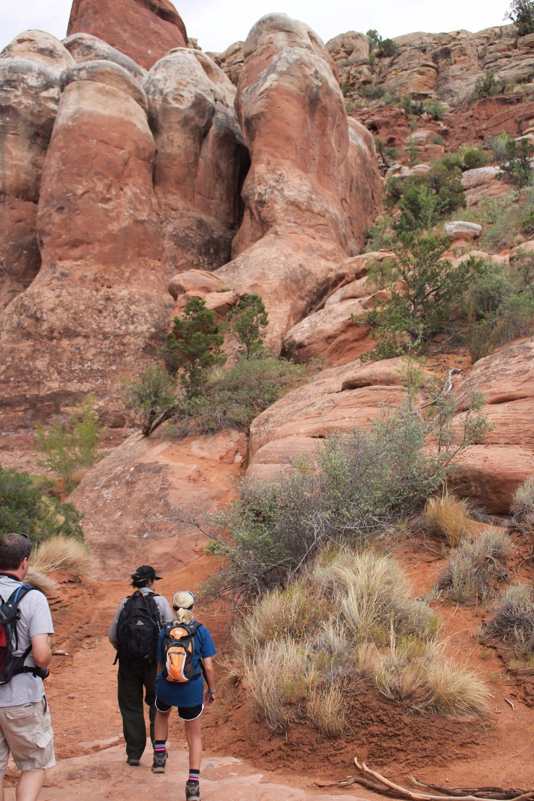 Hikers on a red rock trail beneath tall sandstone formations and desert shrubs.