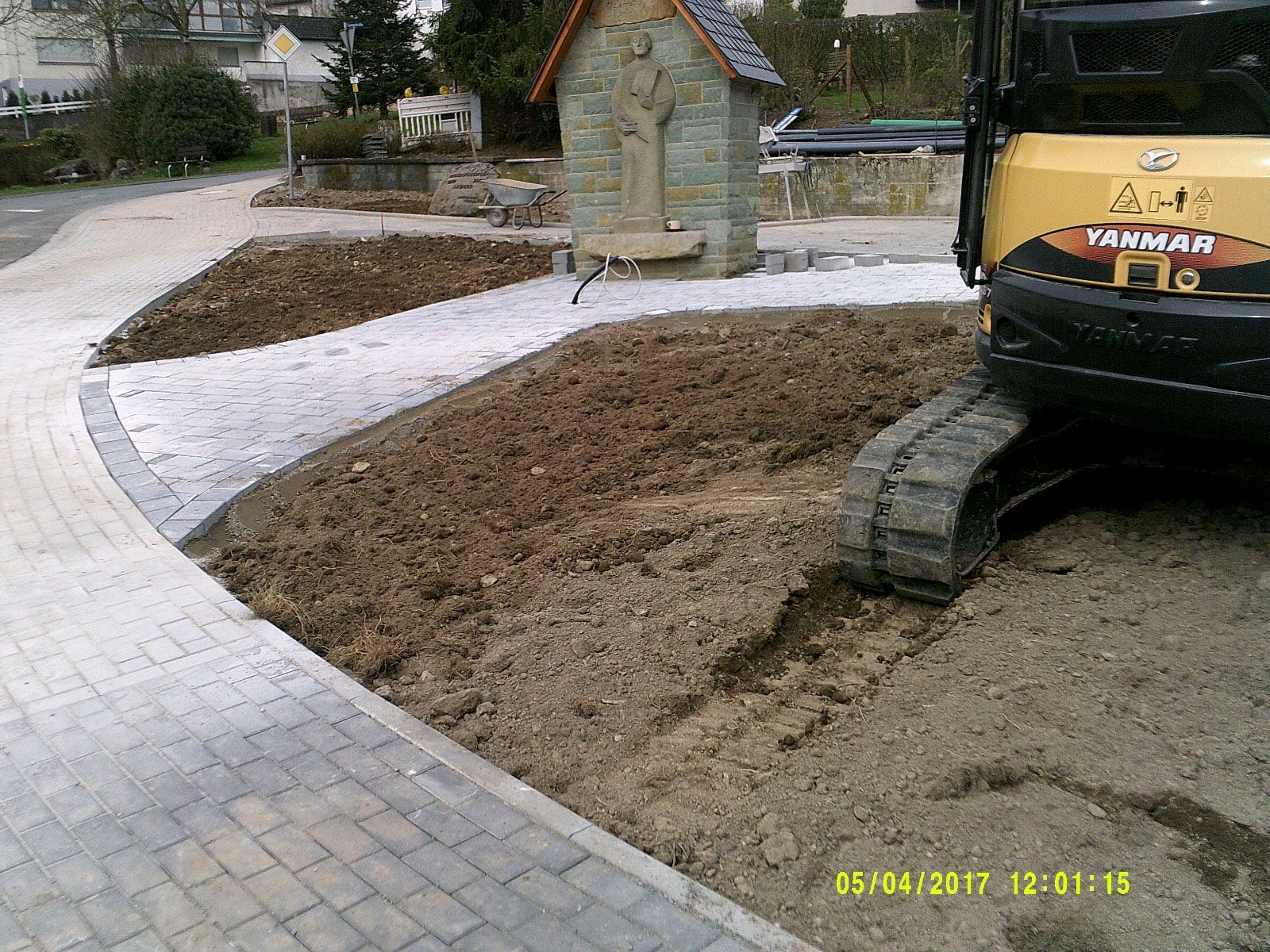 Neben einem gepflasterten Gehweg parkt ein gelber Bagger.