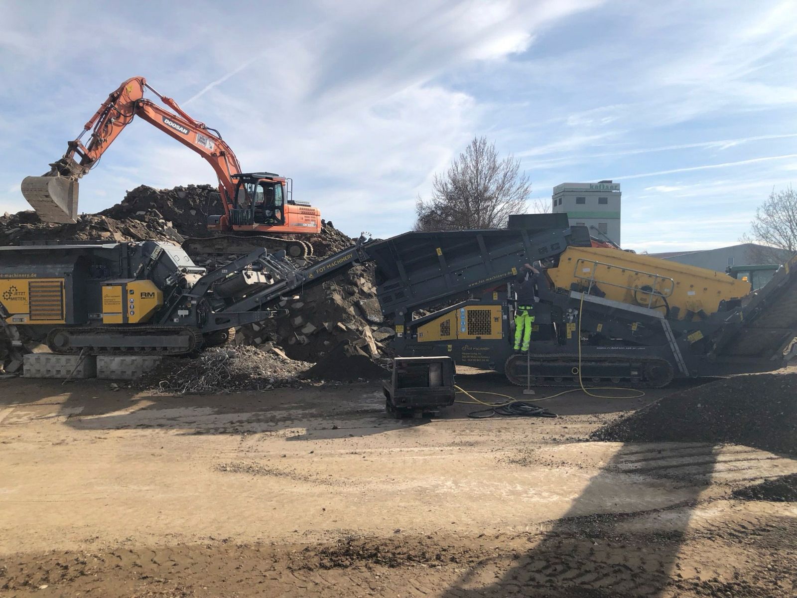 Neben einer großen Maschine gräbt ein großer Bagger einen Erdhaufen.