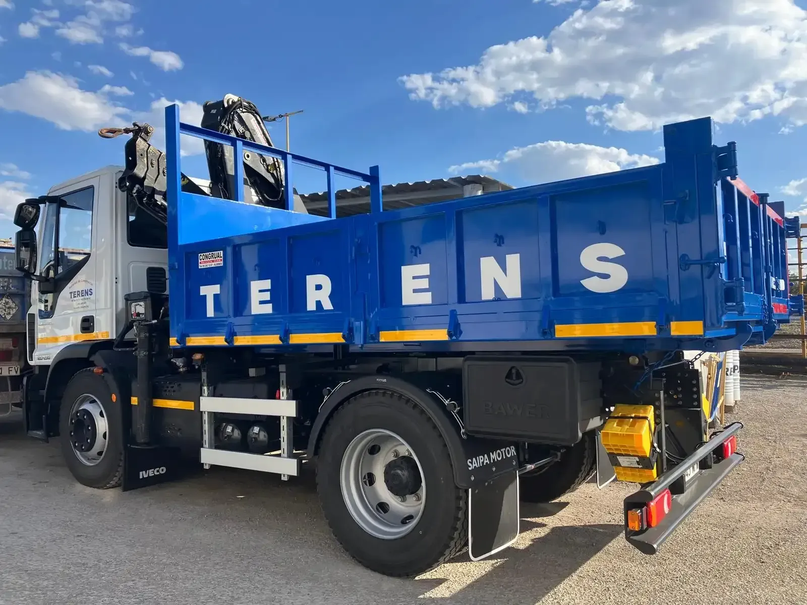 Camión TERENS azul con grúa, estacionado al aire libre contra un cielo nublado.