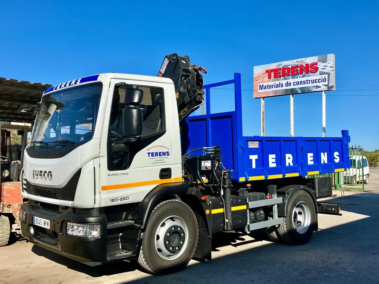 Camión Terens blanco y azul con grúa estacionado al aire libre bajo un cielo azul.