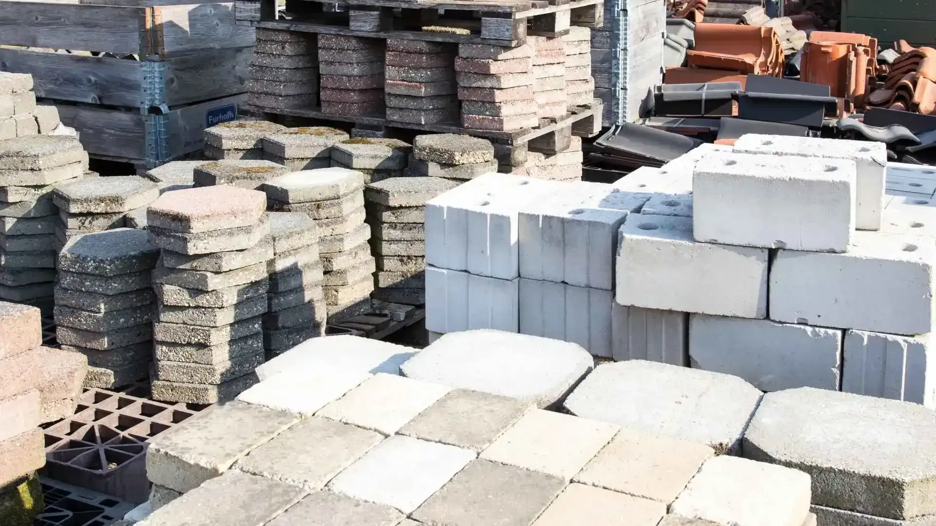 Pilas de diversos adoquines de hormigón y ladrillos en un patio de suministros de construcción al aire libre.