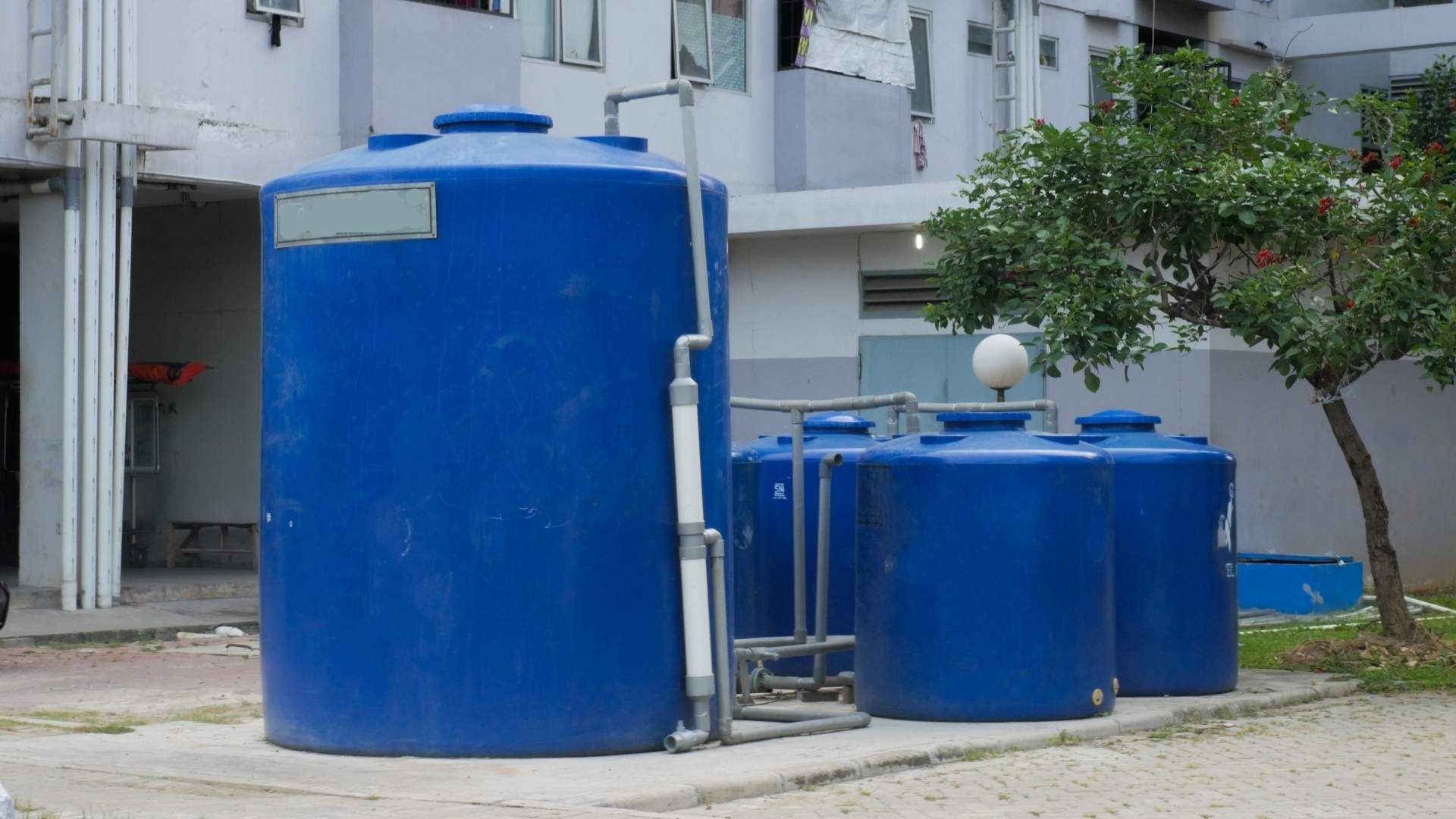 Tanques de agua azules afuera de un edificio, con tuberías de conexión y un pequeño árbol.