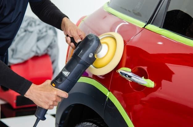 Persona puliendo un coche rojo con una pulidora eléctrica; la cinta verde protege los bordes.