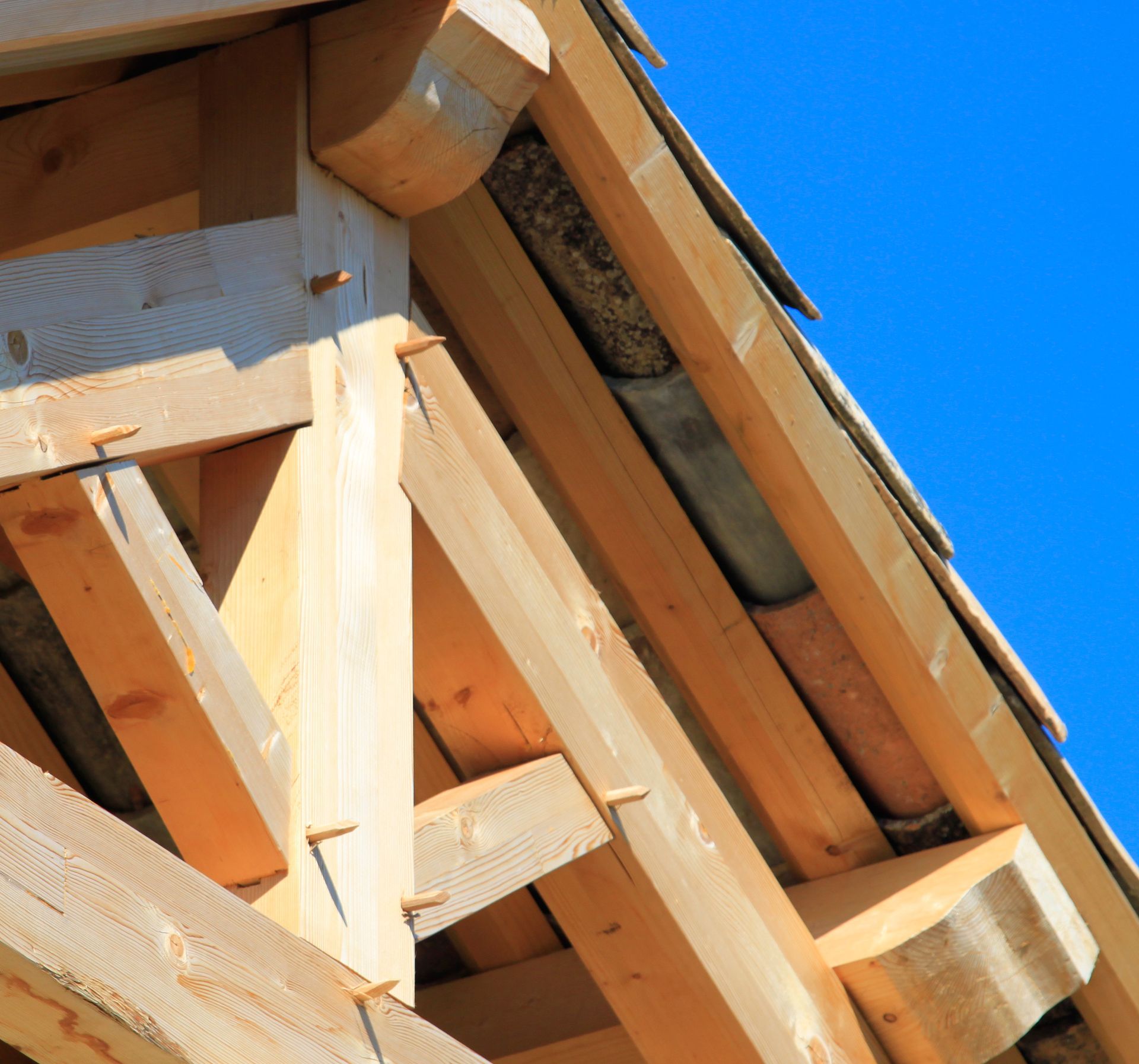 Charpente en bois avec poutres apparentes, joints et matériau sombre sous les bardeaux, sur fond de ciel bleu clair.