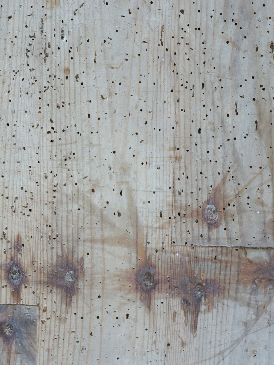 Panneau de bois usé présentant de nombreux petits trous sombres et des têtes de boulons rouillées.
