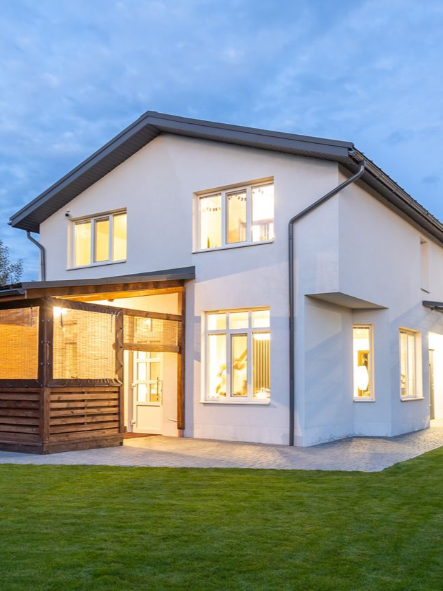 Maison blanche à deux étages avec terrasse en bois et pelouse verte au crépuscule.