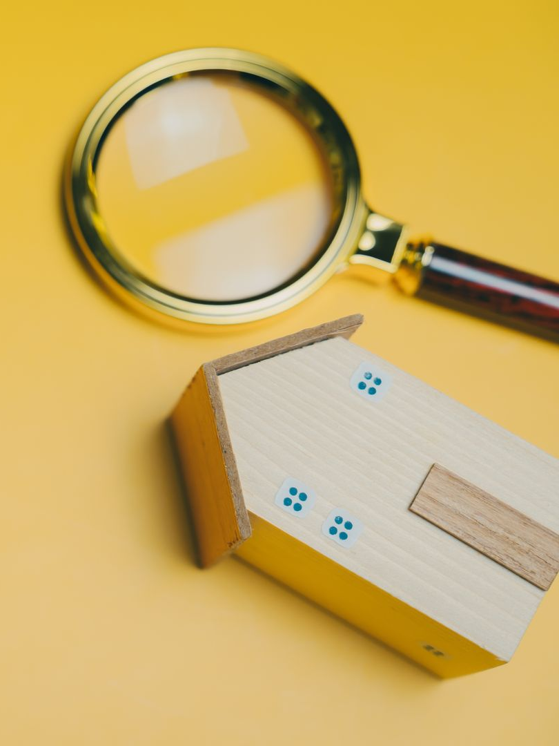Loupe placée au-dessus d'une maquette de maison en bois sur fond jaune, symbolisant l'inspection du logement.