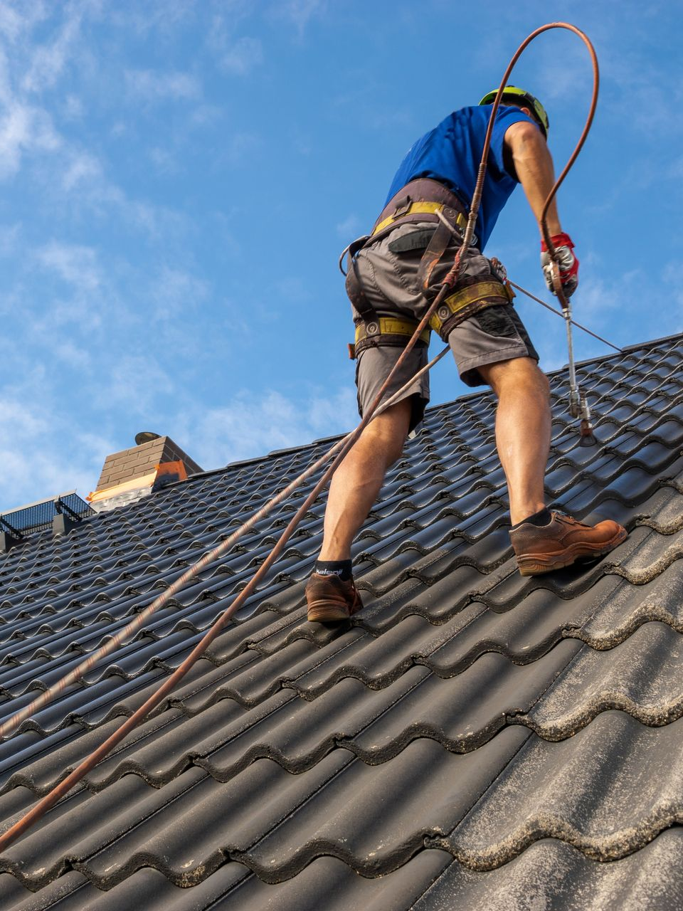 Un couvreur, harnaché et en sécurité, travaille sur un toit de tuiles sombres se détachant sur un ciel bleu.