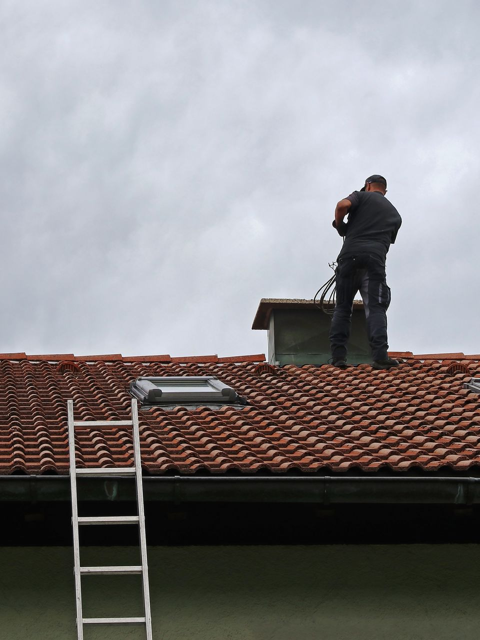 Une personne sur un toit de tuiles rouges, près d'une cheminée, utilisant des outils, avec une échelle et un ciel couvert.