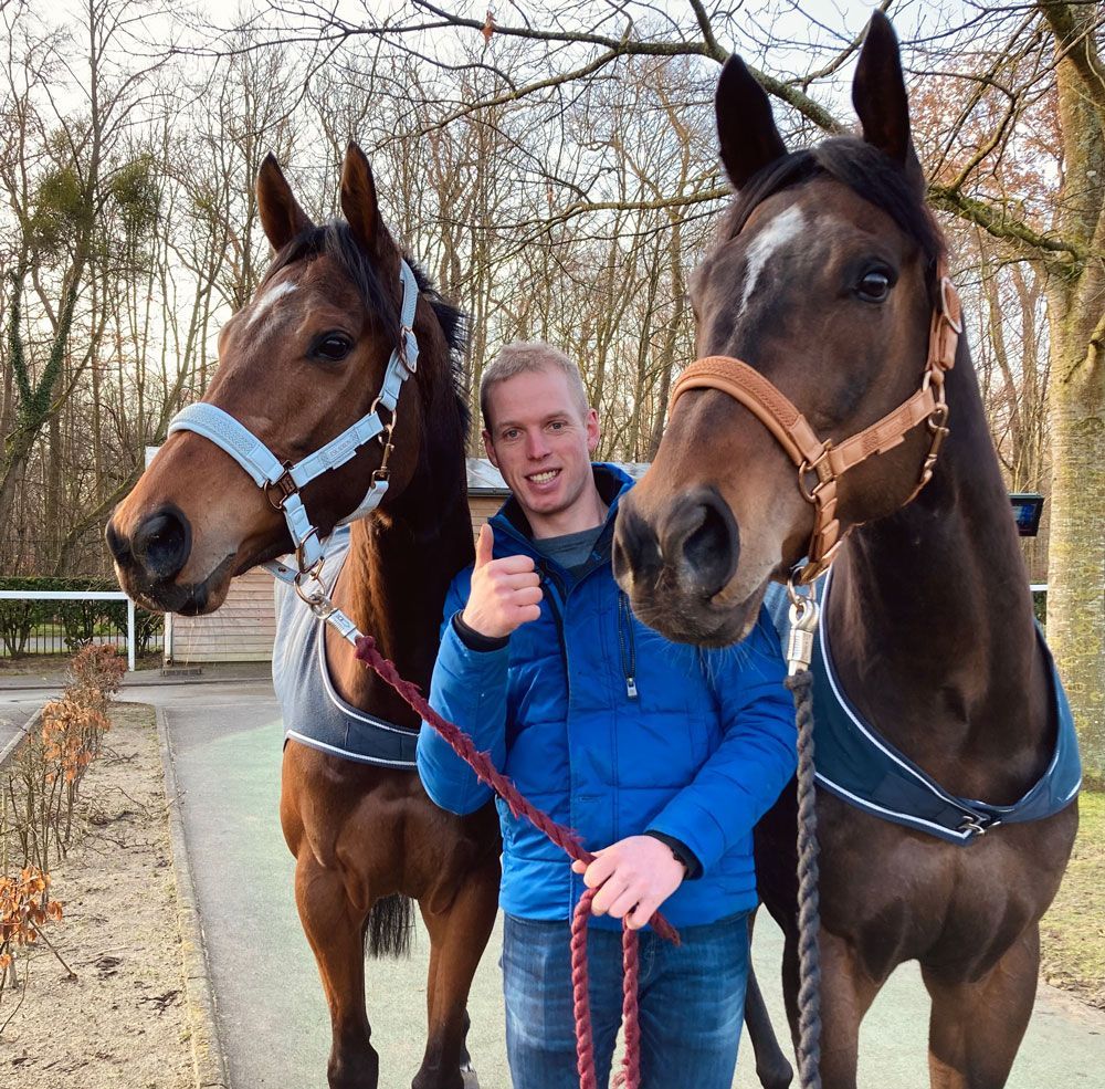 Ein Mann, der den Daumen nach oben streckt, hält die Zügel eines Pferdes und steht im Freien neben zwei braunen Pferden.