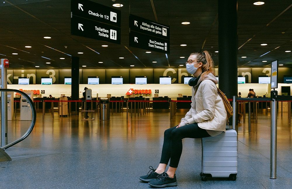 Una mujer que lleva una máscara está sentada sobre una maleta en un aeropuerto.