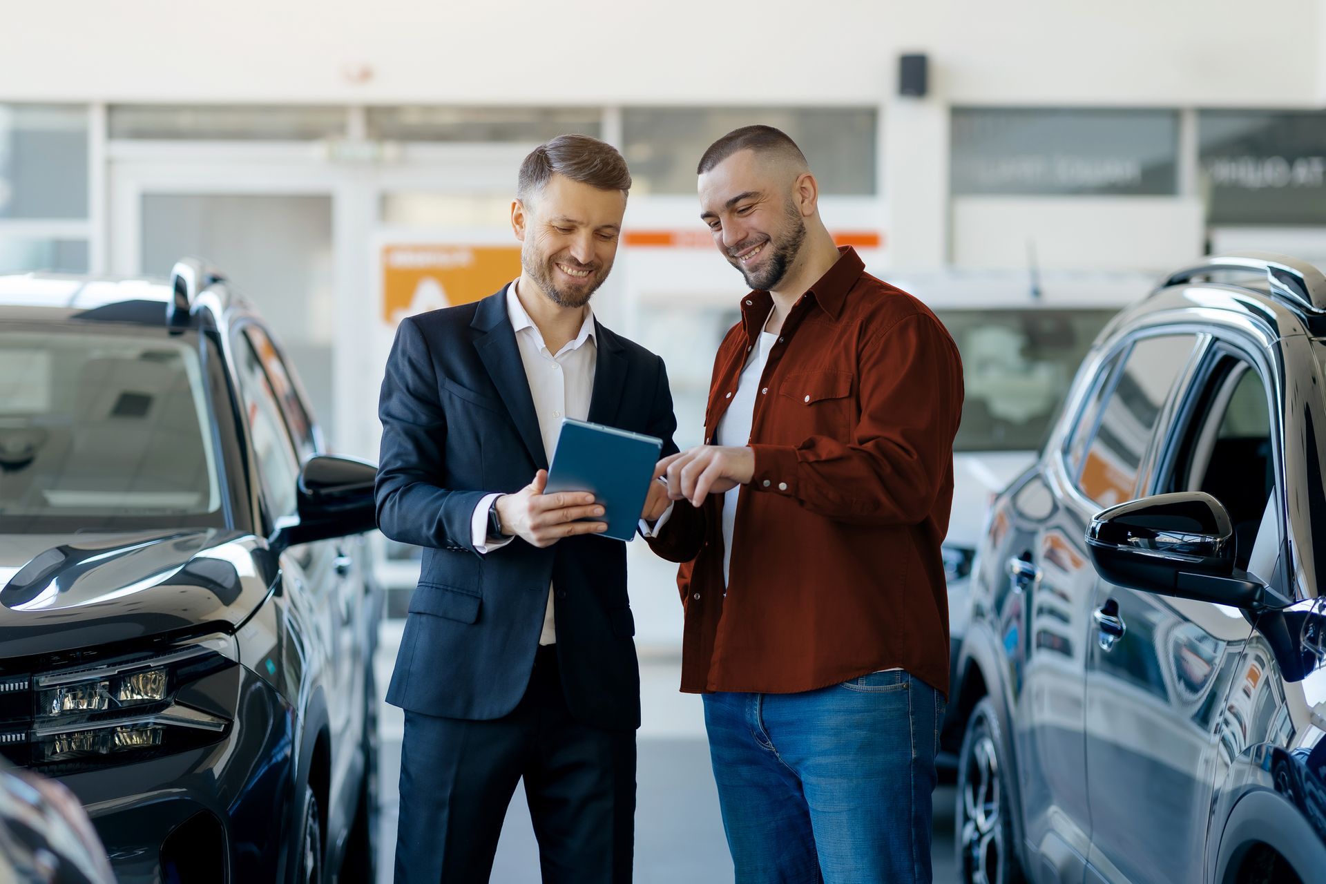 Dans une concession automobile, un vendeur en costume présente une tablette à un client, entre deux véhicules.