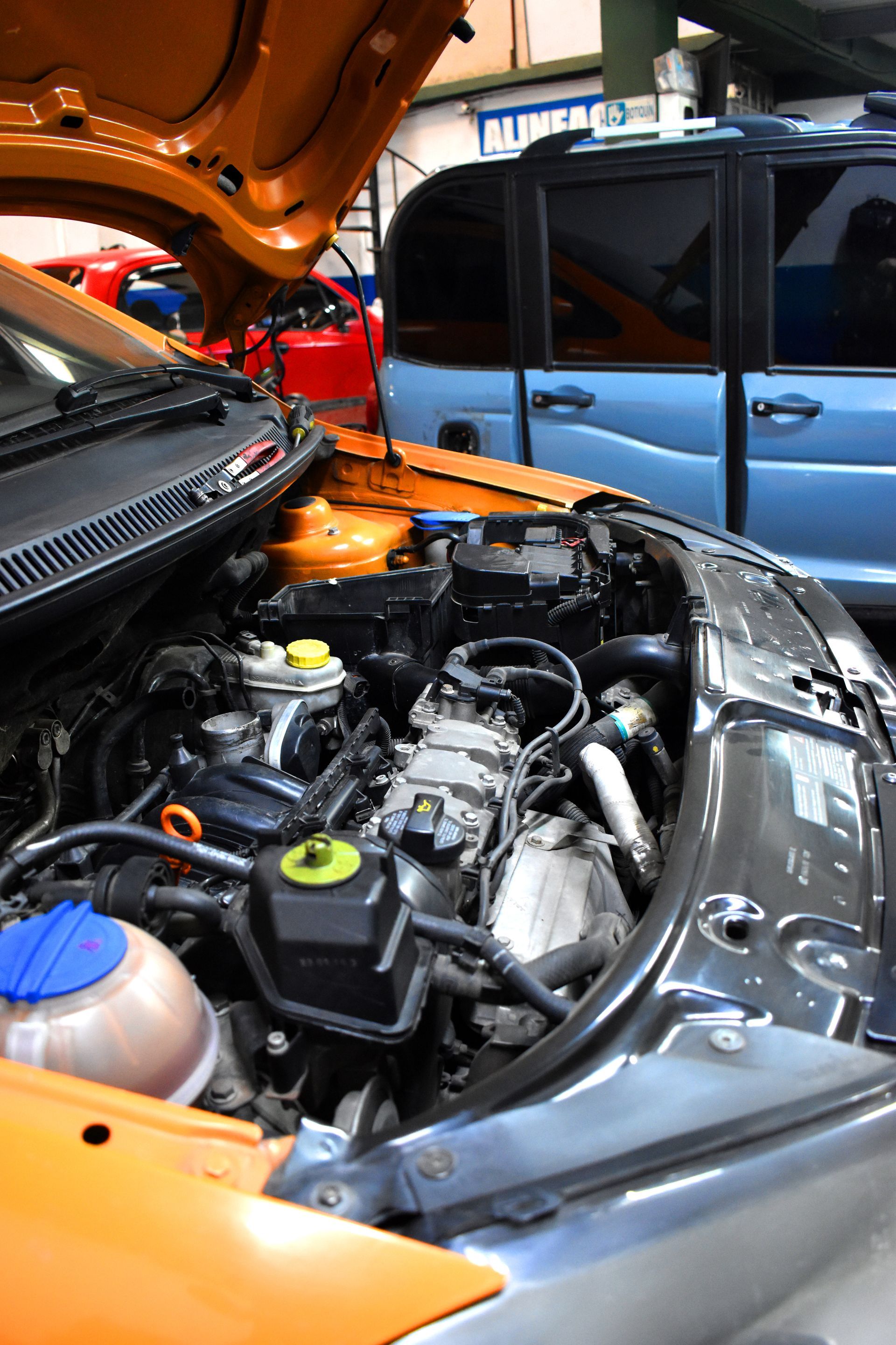Gros plan sur le compartiment moteur ouvert d'une voiture orange dans un atelier, avec un véhicule bleu clair.