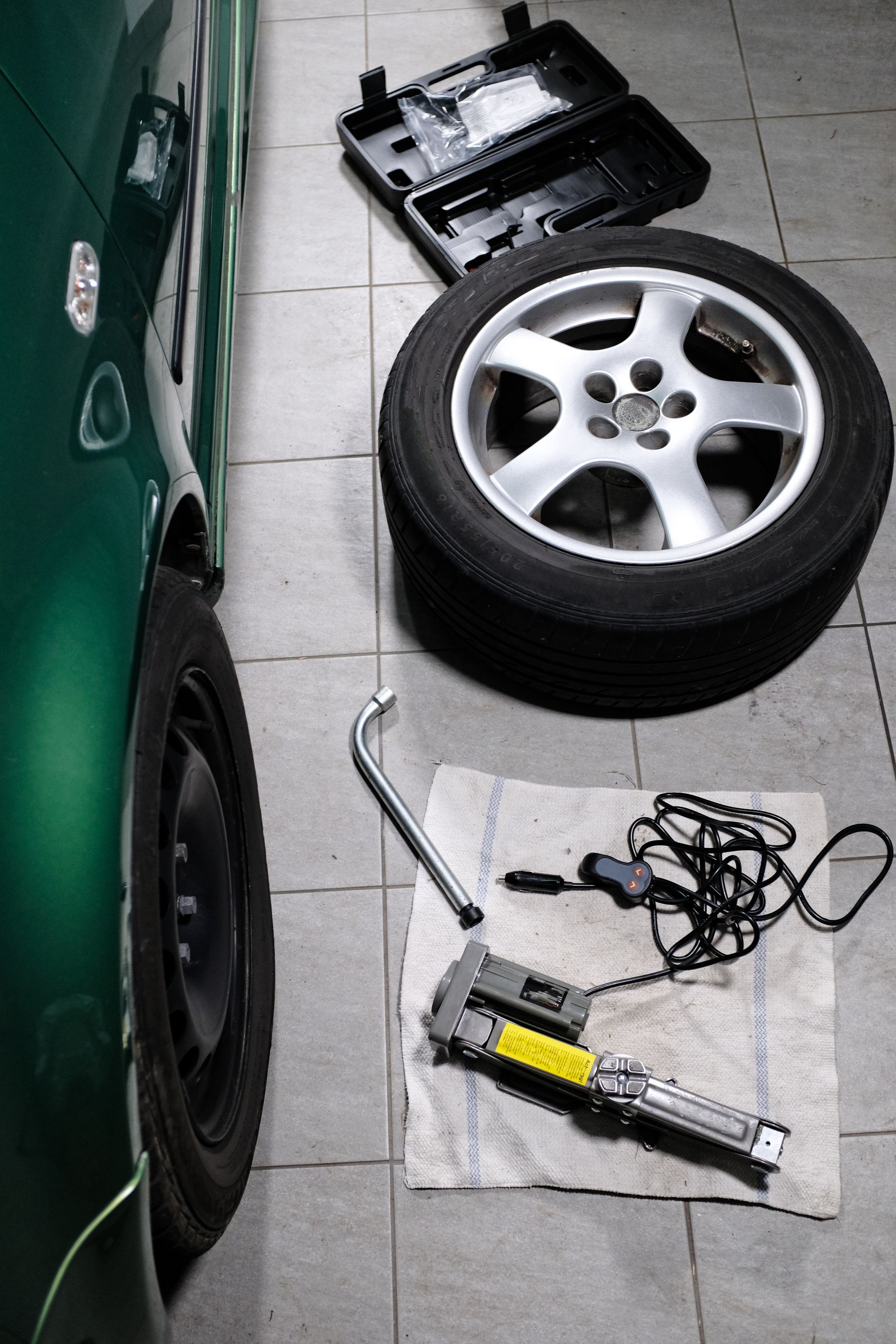 Une voiture, une roue de secours, une clé à roue et un cric électrique disposés sur un carrelage pour un changement de pneu.