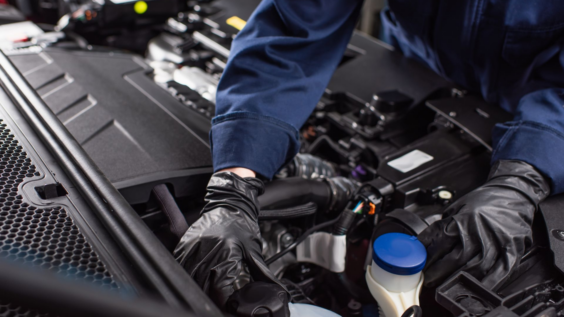 Un mécanicien vêtu d'une combinaison bleue et de gants noirs effectue l'entretien d'un moteur de voiture.