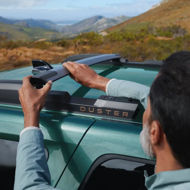 Une personne ajuste les barres de toit modulaires d'une Dacia Duster verte sur fond de paysage montagneux.