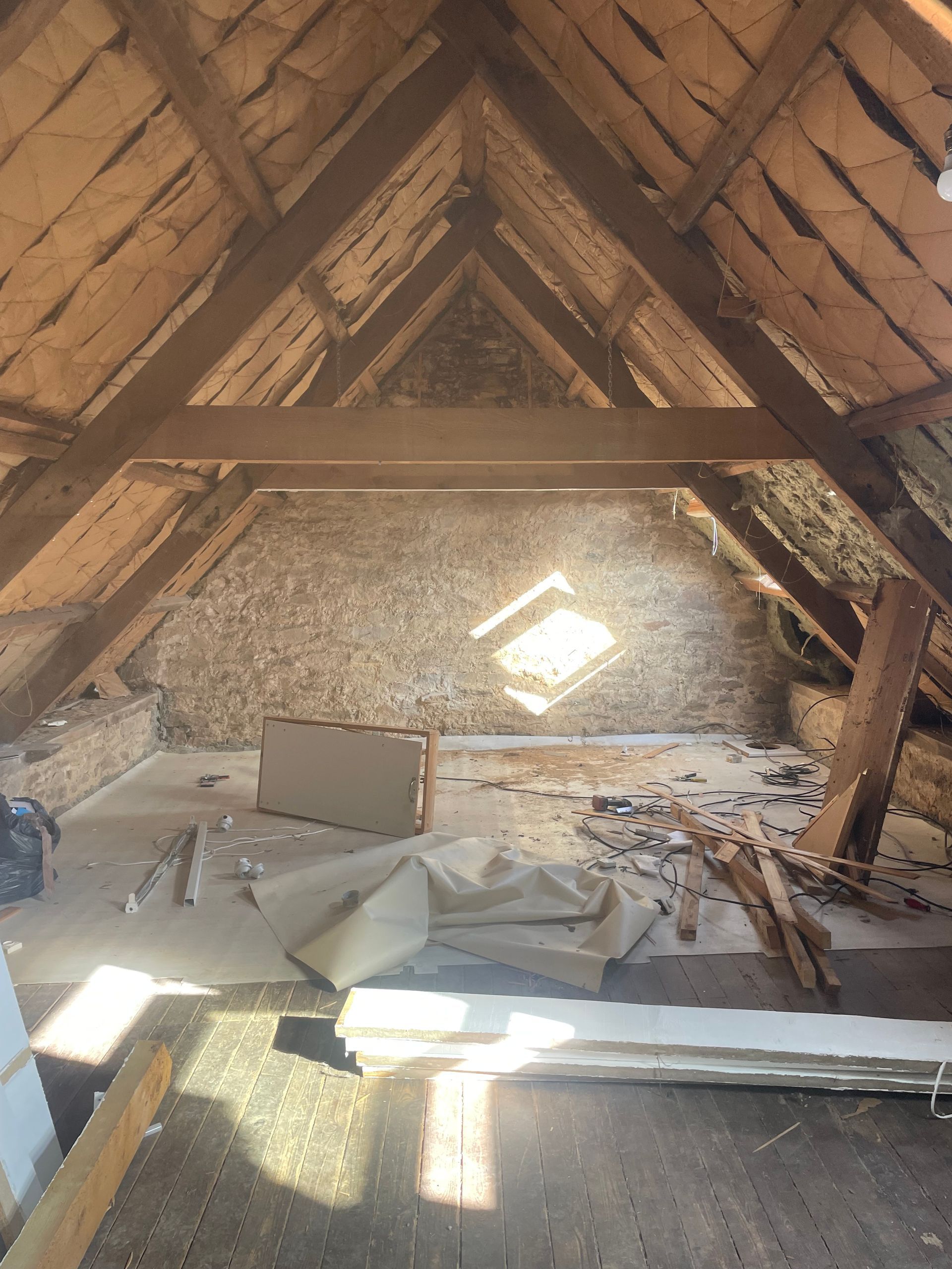 Grenier avec poutres apparentes, mur en pierre et petite lucarne. Des matériaux de construction sont éparpillés sur le sol.