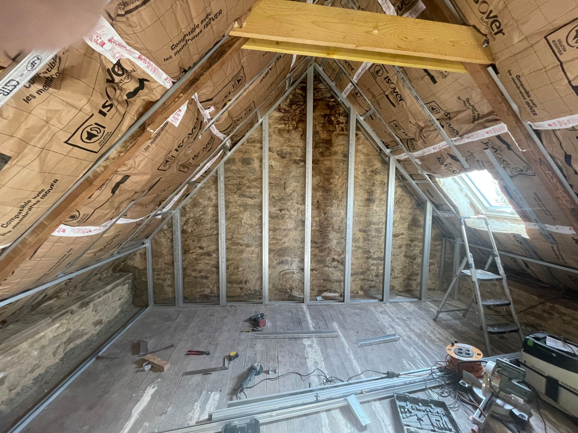 Intérieur du grenier isolé, avec ossature métallique et chevrons en bois. Outils et matériaux au sol.
