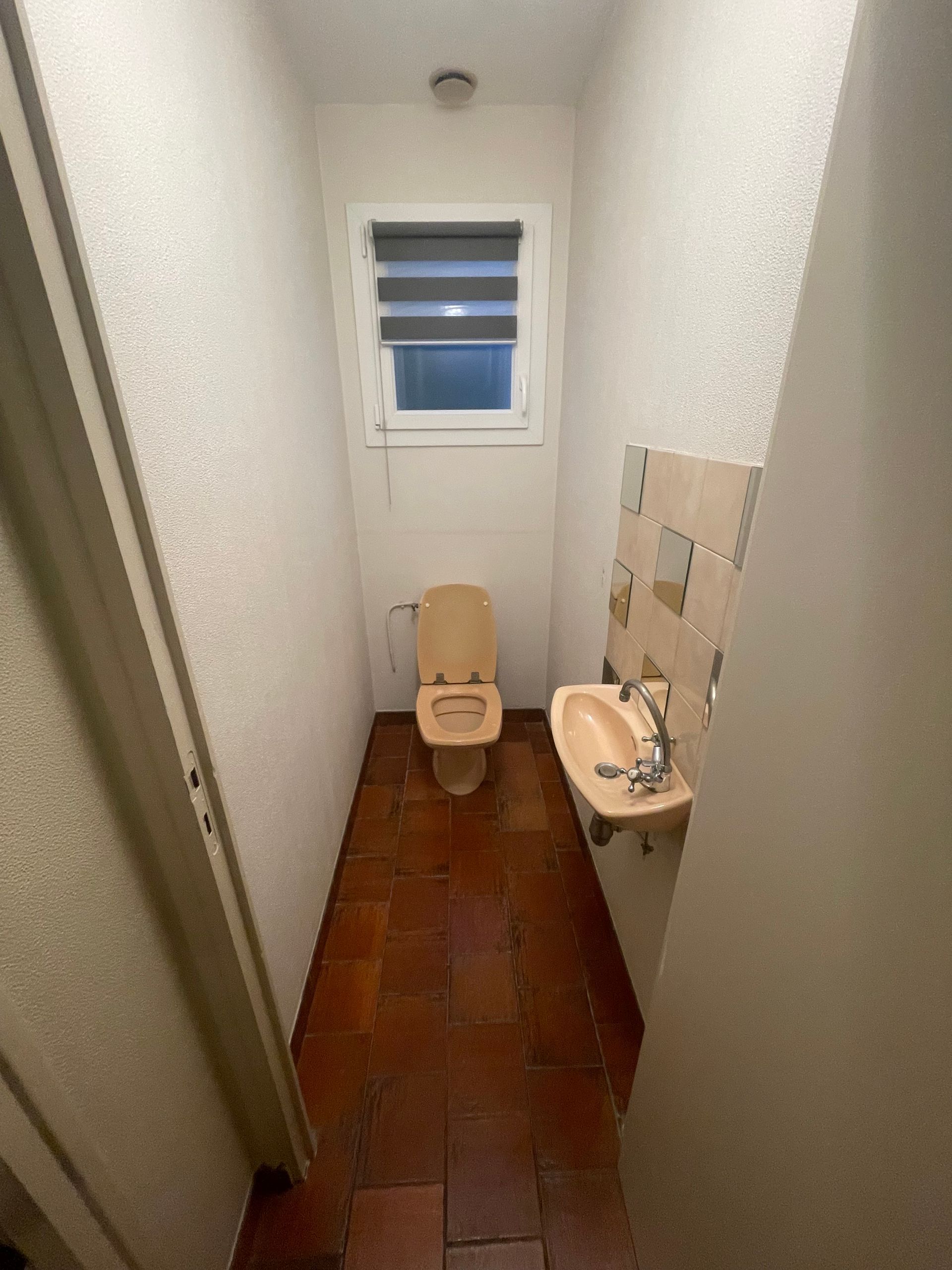 Petite salle de bains avec toilettes, lavabo et petite fenêtre ; sol carrelé marron et murs blancs texturés.