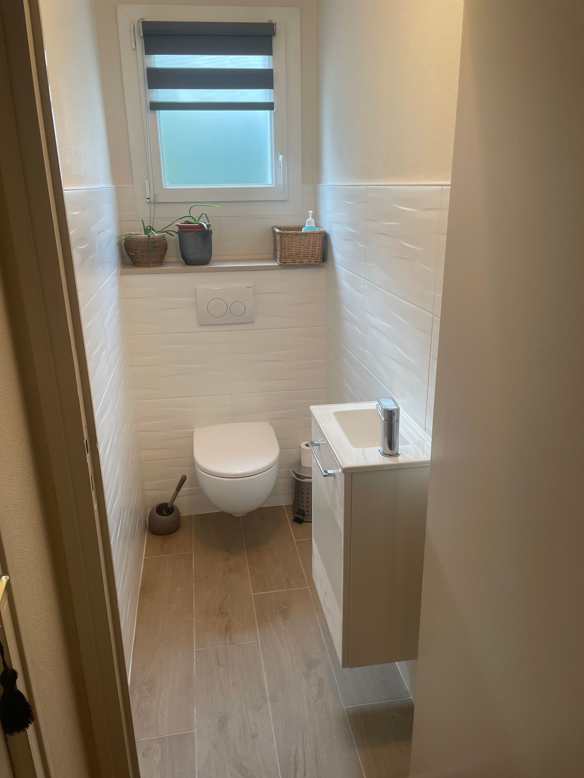 Petite salle de bains aux murs blancs, lavabo suspendu et toilettes ; une fenêtre et des plantes se trouvent au-dessus des toilettes.