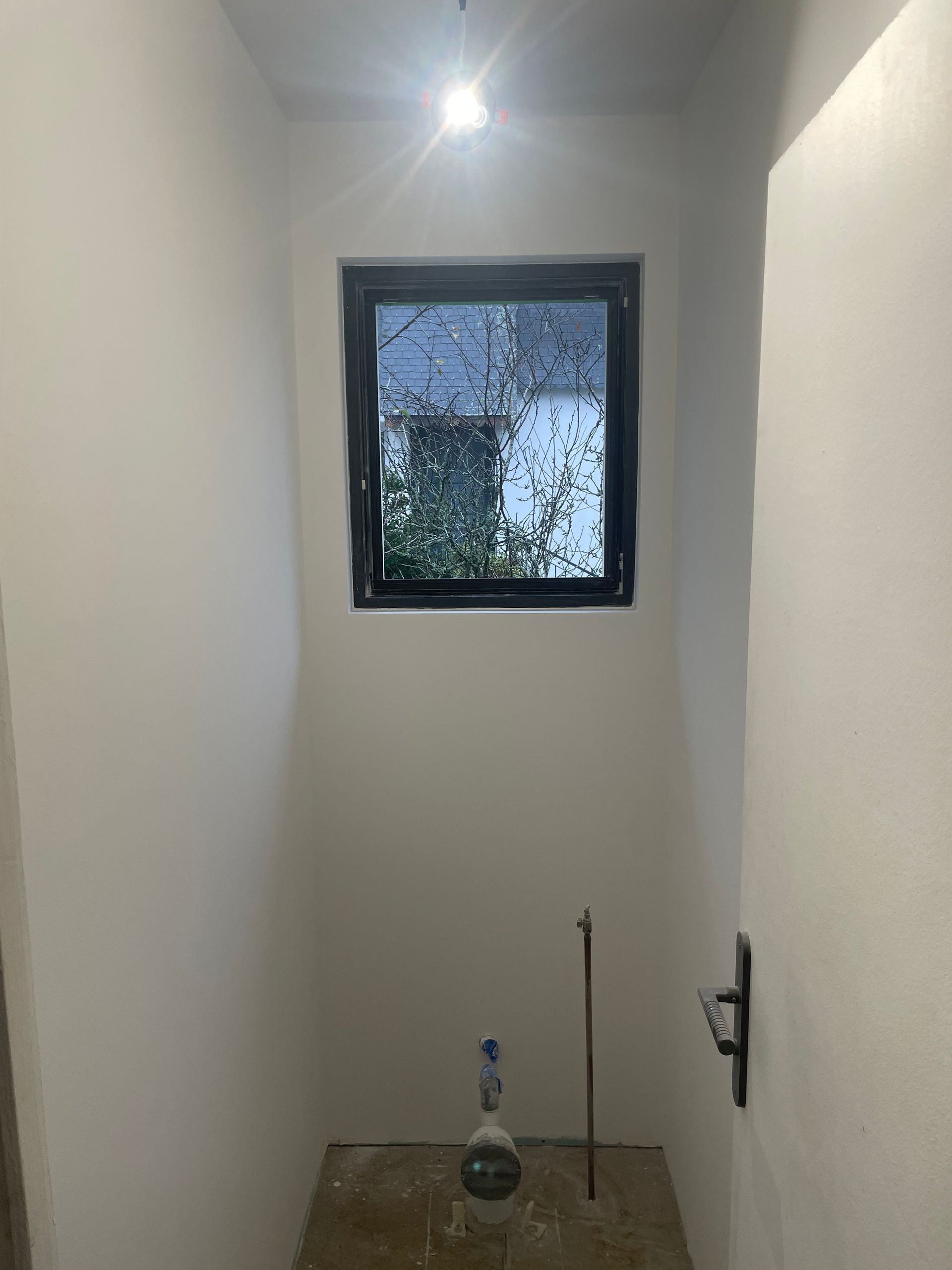 Petite pièce inachevée avec une fenêtre. Murs blancs, cadre de fenêtre sombre, installations sanitaires apparentes.