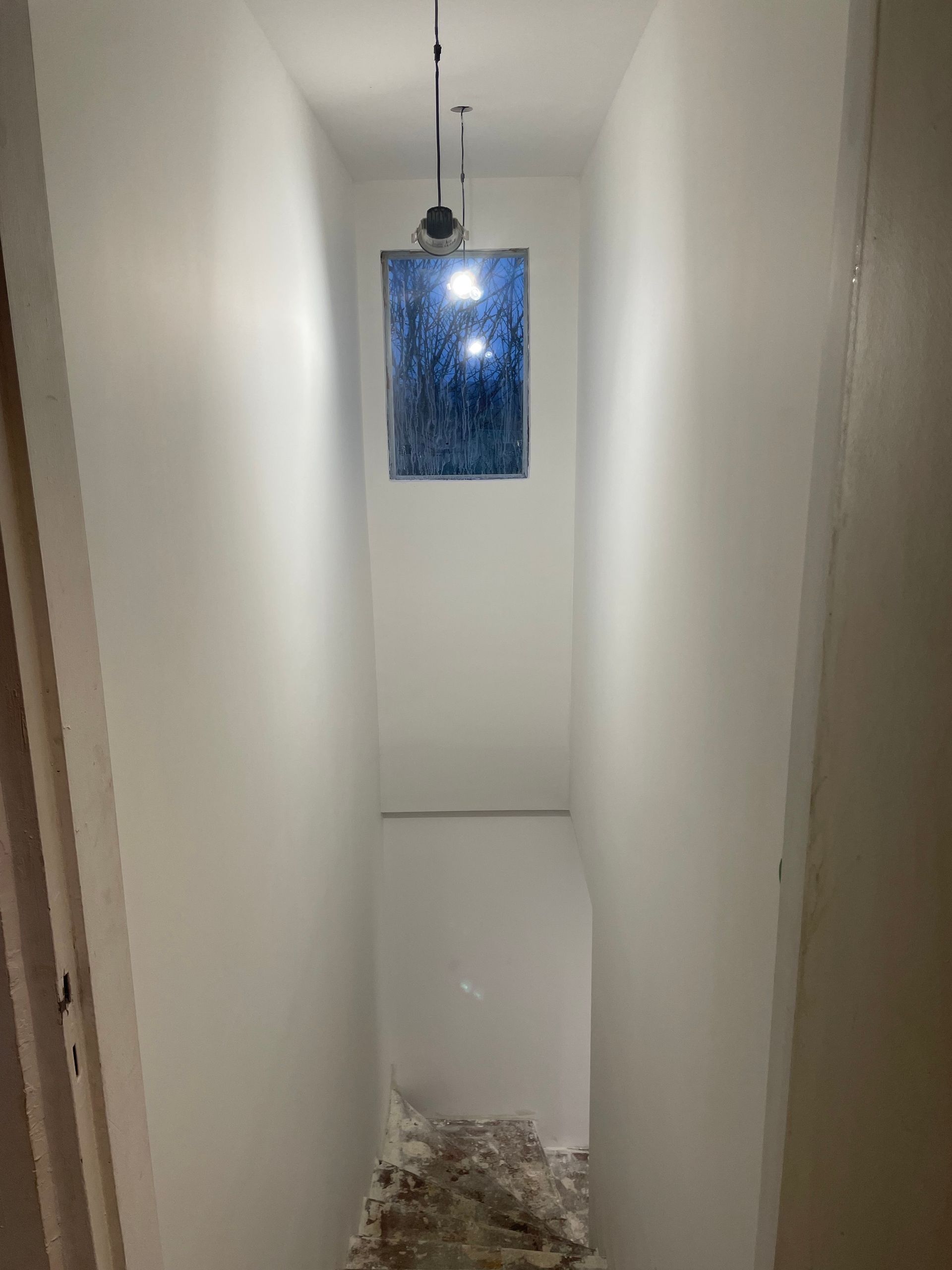Couloir étroit aux murs blancs et une petite fenêtre au fond. Débris au sol.