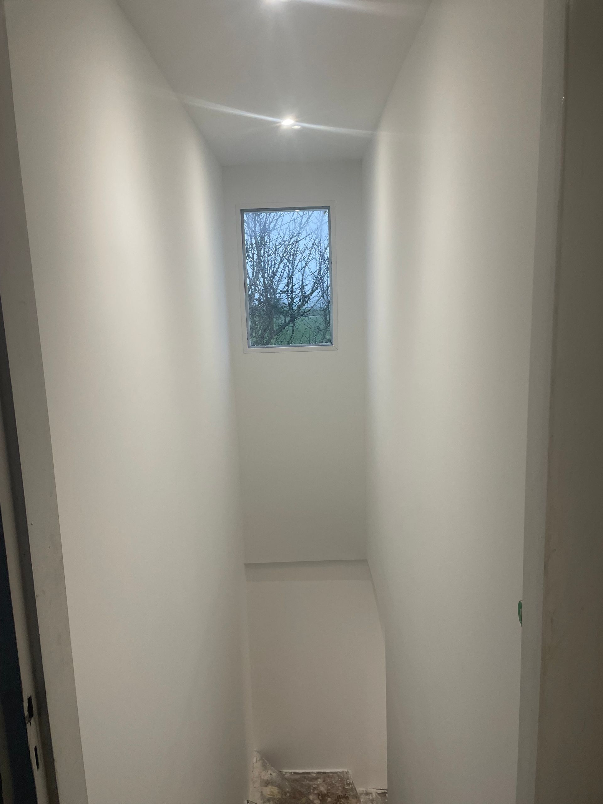 Couloir blanc étroit avec une petite fenêtre au fond, menant à un escalier.