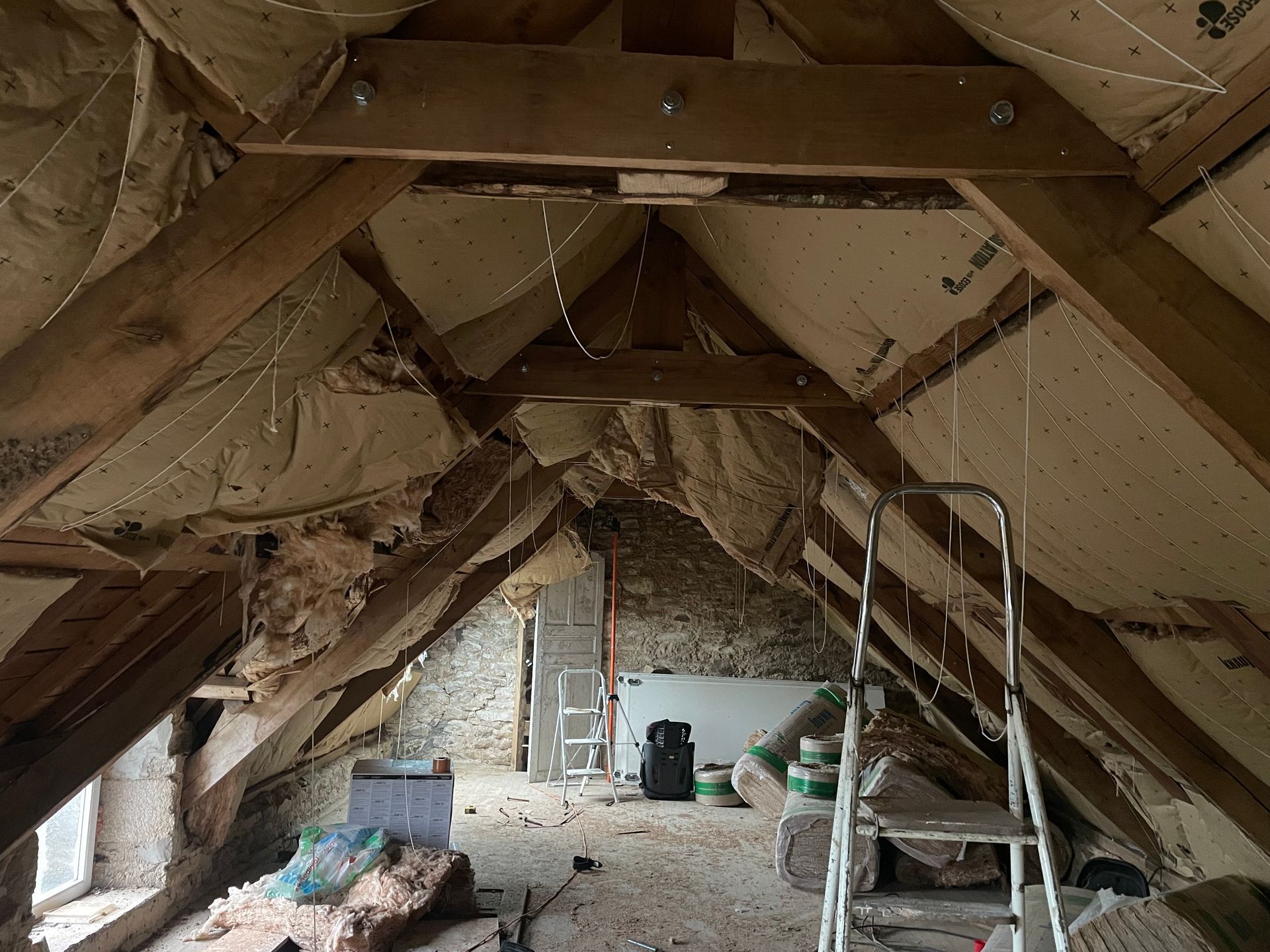Intérieur du grenier avec poutres en bois apparentes, isolation et matériaux de construction ; une échelle est visible.