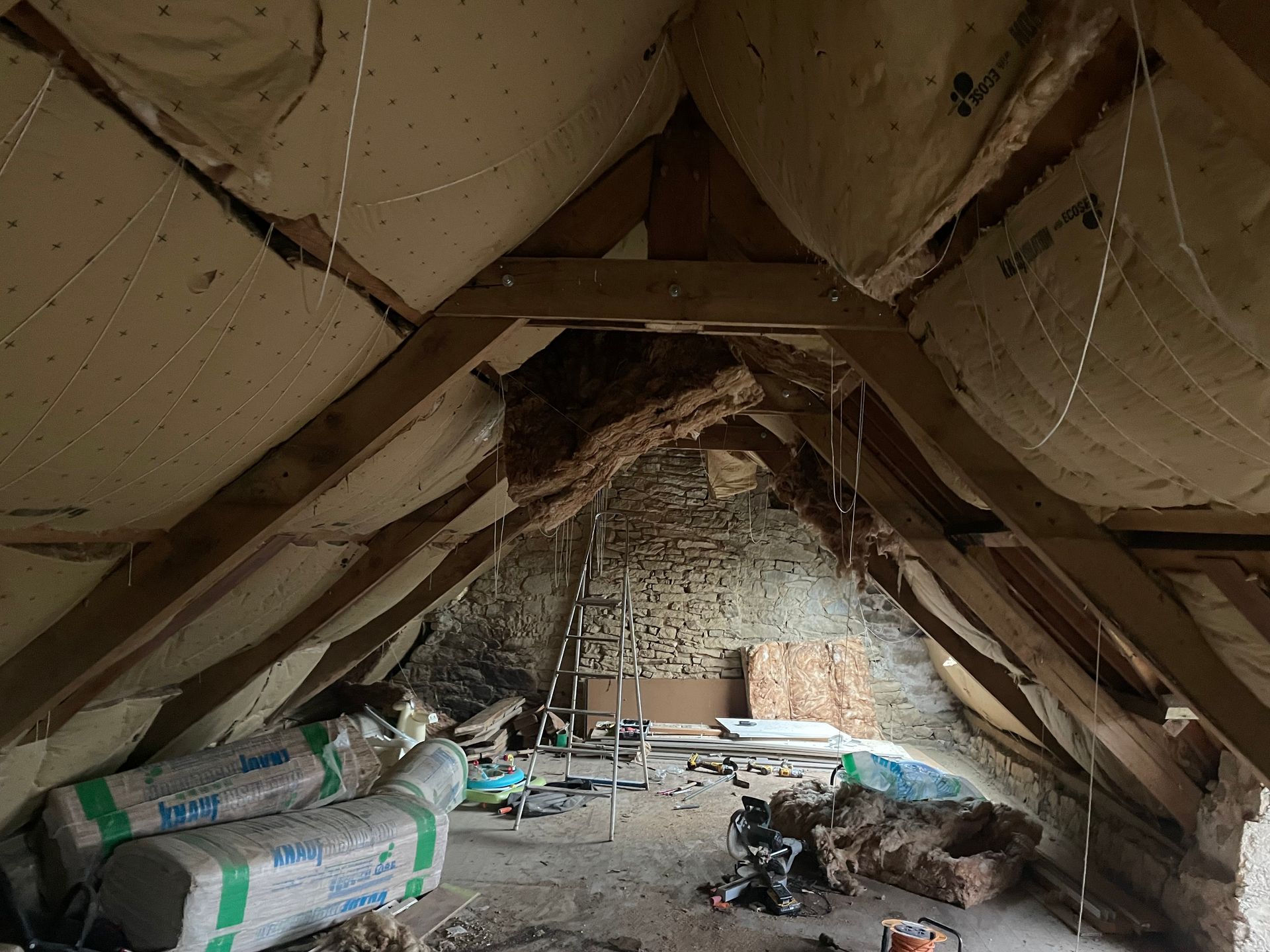 Intérieur d'un grenier avec poutres en bois, isolation et échelle, possiblement en cours de rénovation.