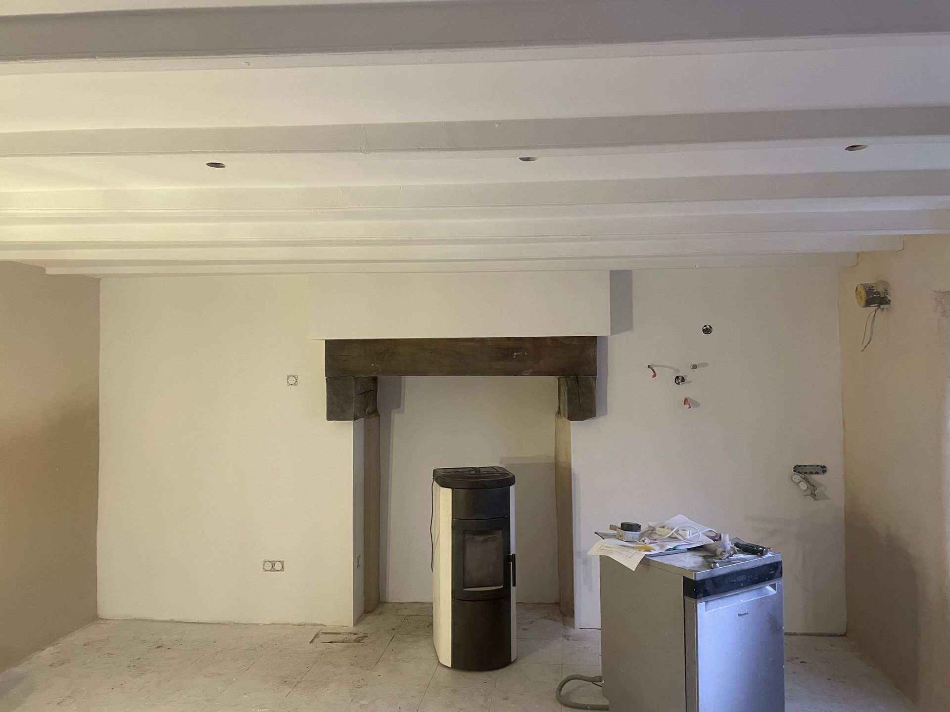 Intérieur d'une pièce en rénovation avec cheminée, poutres apparentes et poêle. Murs et poutres de plafond de couleur claire.