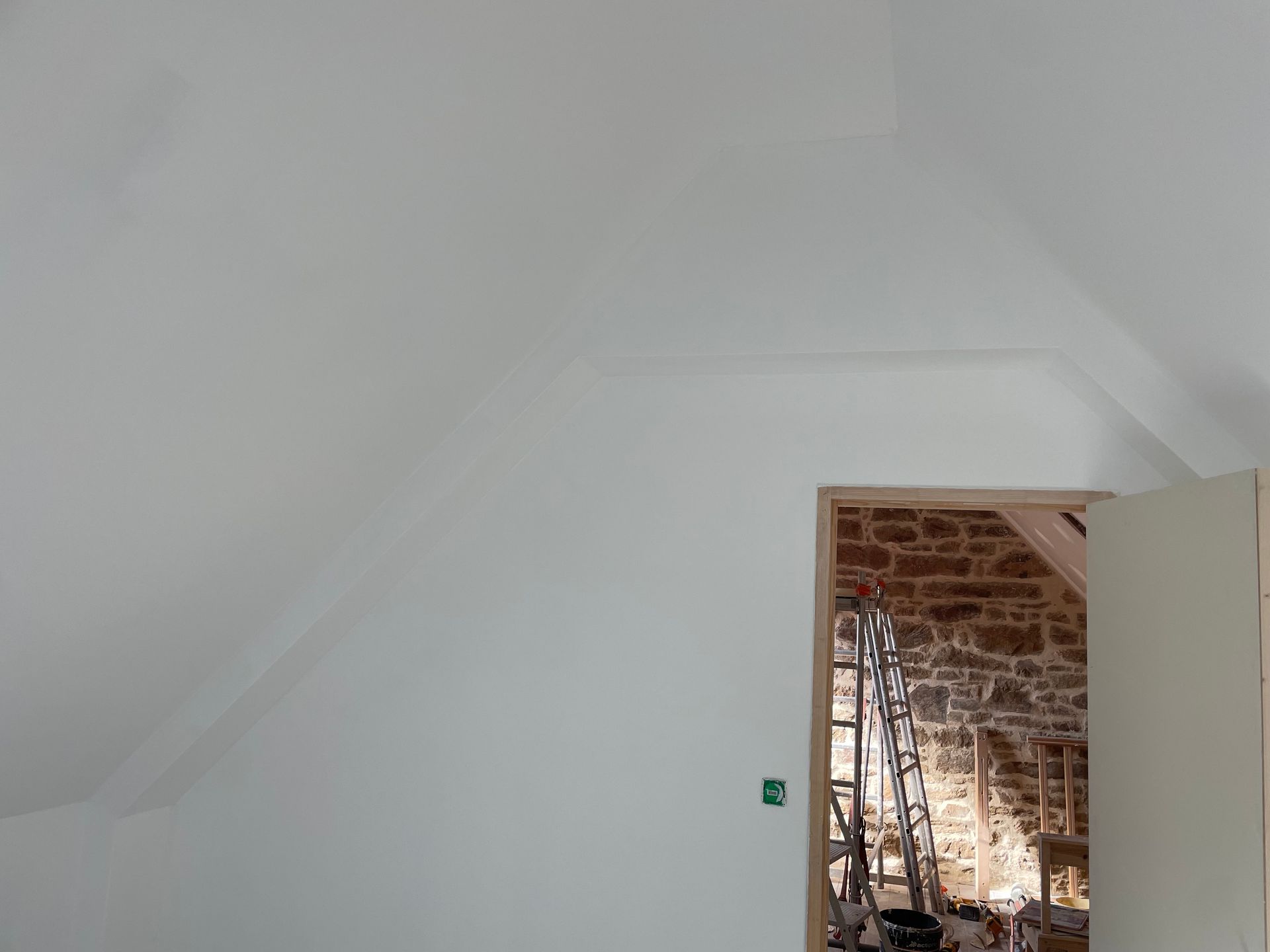 Pièce peinte en blanc avec plafond en pente et porte donnant sur un espace aux murs de pierre.