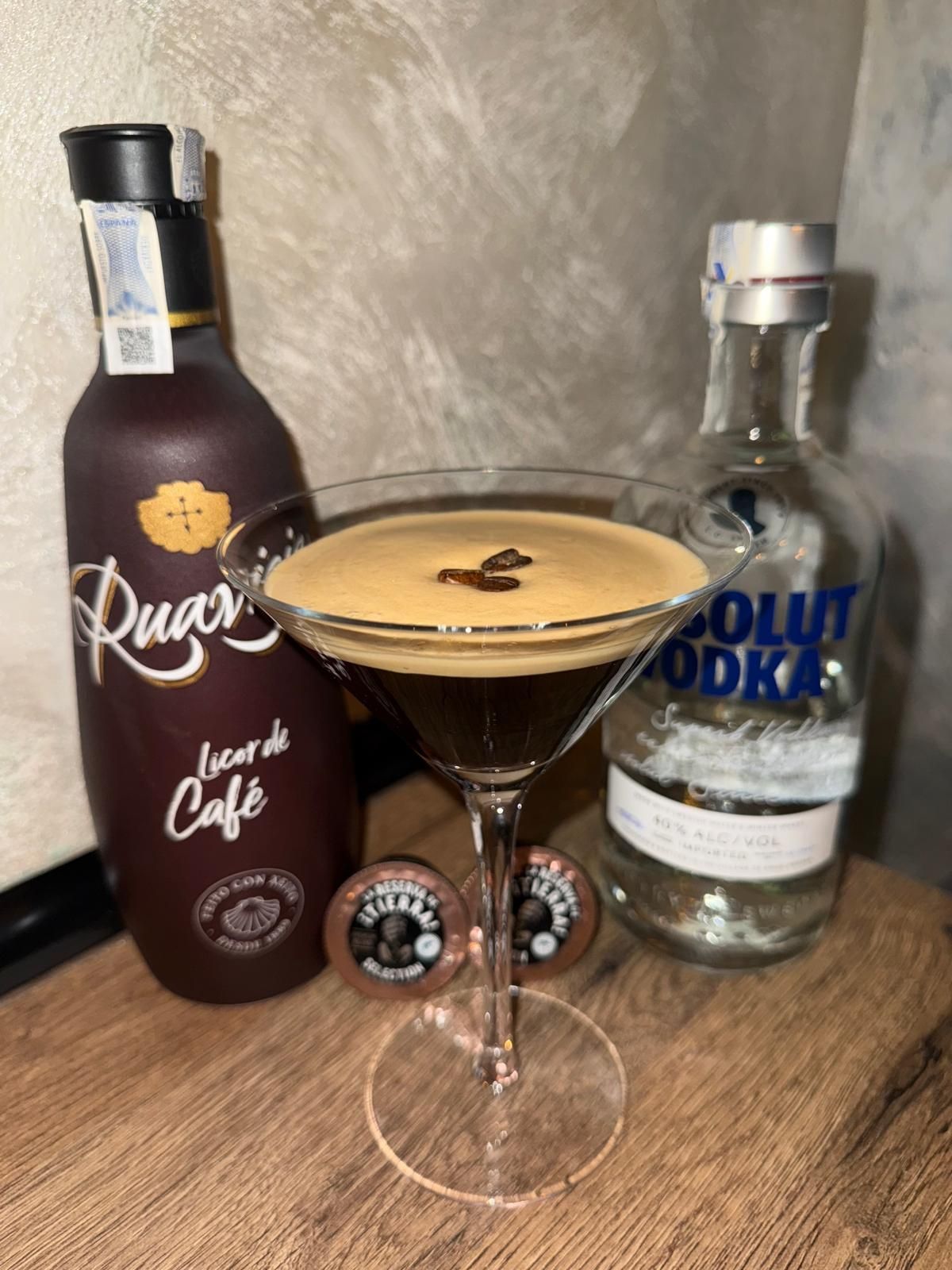 Espresso Martini en copa de tallo con botellas de licor de café y vodka sobre una superficie de madera.