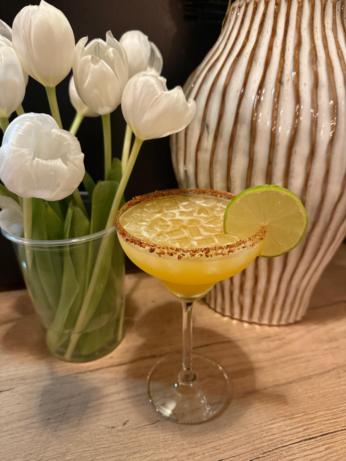Cóctel con lima y sal se encuentra sobre una superficie de madera junto a tulipanes blancos en un jarrón y un jarrón a rayas.