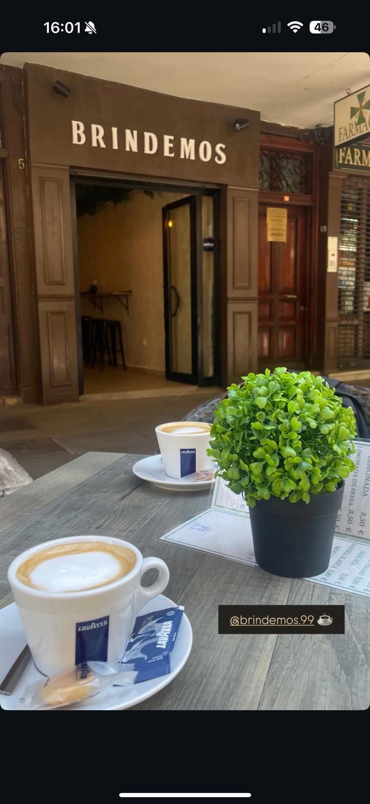 Tazas de café sobre una mesa, con una planta verde, frente a la entrada de un café que dice 