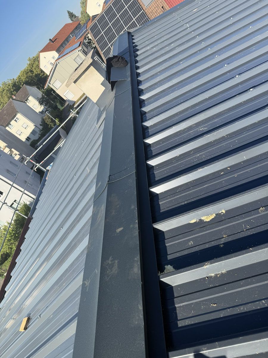 Blick von oben auf den metallenen First eines dunkelgrauen Rippendachs an einem sonnigen Tag.