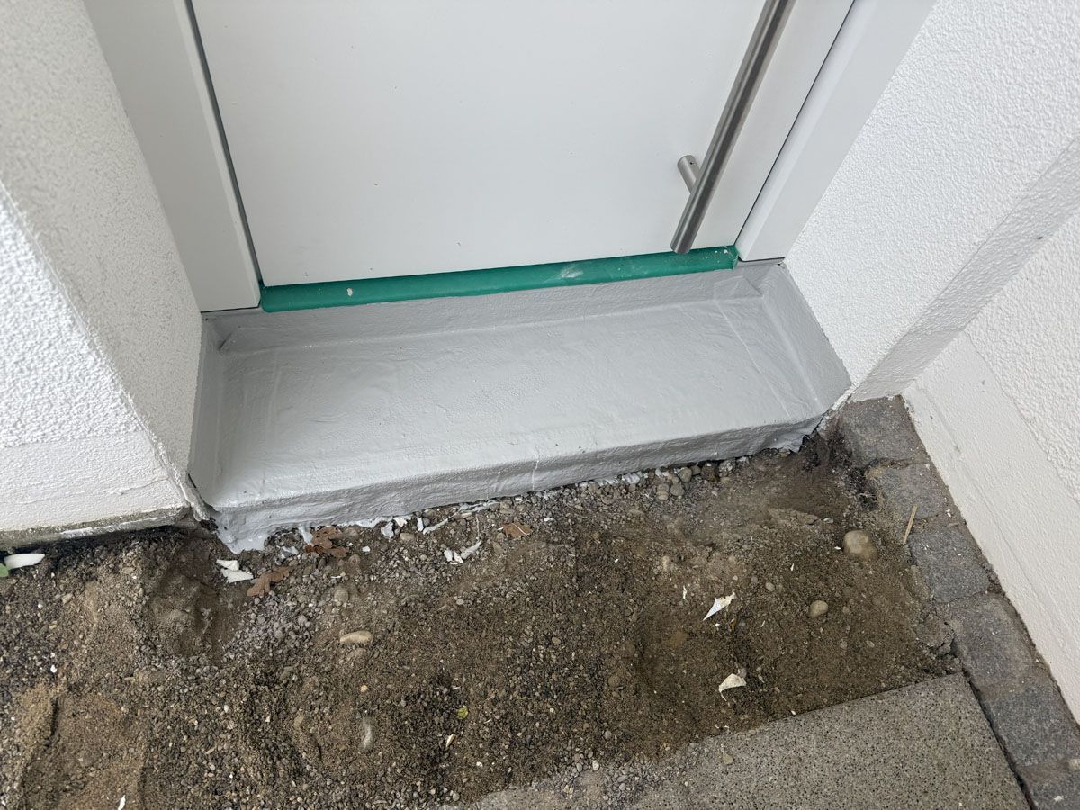 Eine weiße Türschwelle mit einer grauen Betonstufe, die zu einem unbefestigten Erdboden in der Nähe einer Außenwand des Gebäudes führt.