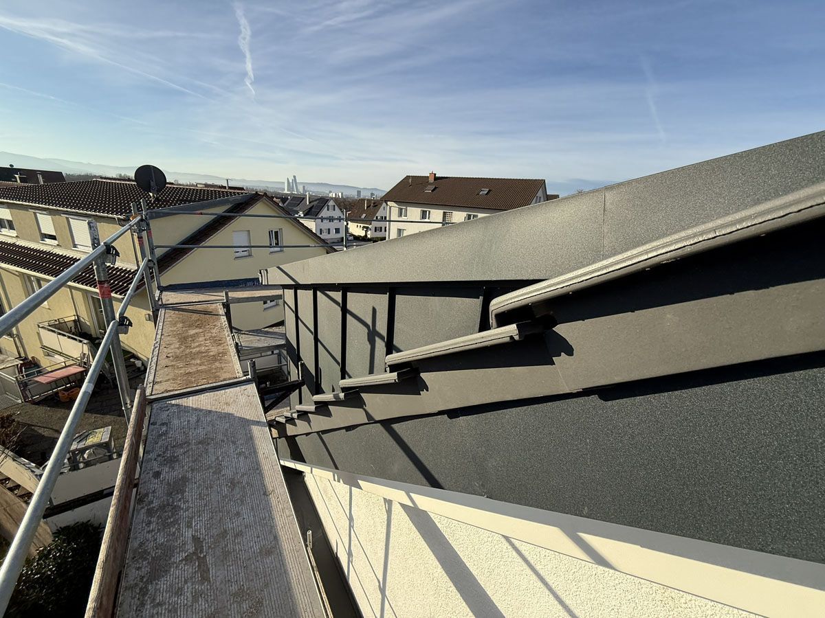 Blick von einem Baugerüst über ein dunkel geziegeltes Dach und nahegelegene Wohnhäuser unter blauem Himmel.