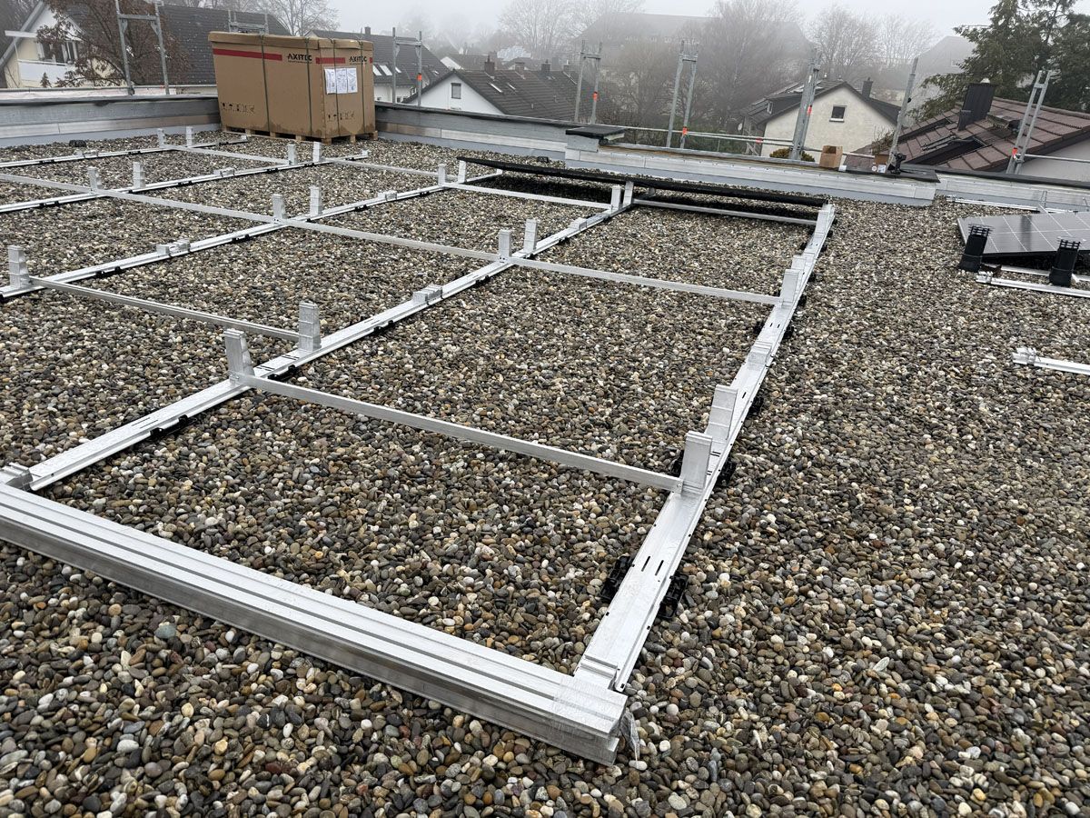 Auf einem flachen, mit Kies bedeckten Dach wird ein metallener Montagerahmen für Solarpaneele installiert.