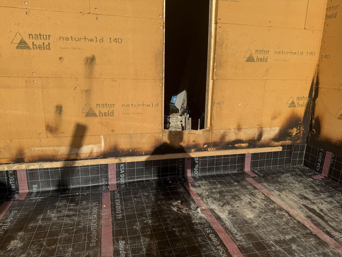 Baustelle mit orangefarbenen Wandplatten und schwarzem, wasserdichtem Membranboden, die eine zentrale, türähnliche Öffnung aufweist.