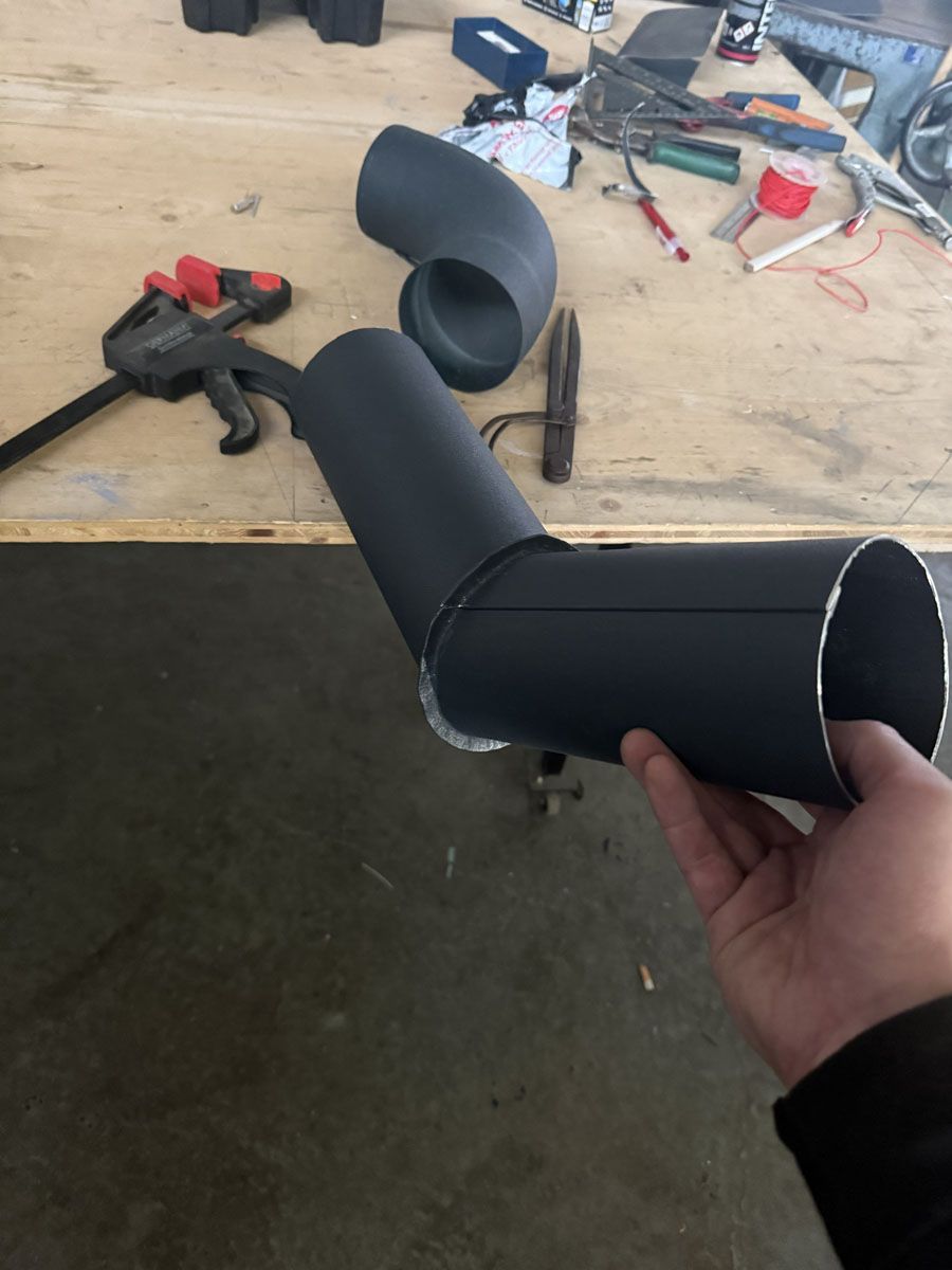 Eine Hand hält zwei miteinander verbundene, mattschwarze Rohrsegmente über einem Werkstatttisch mit Werkzeugen und einer Klemme.