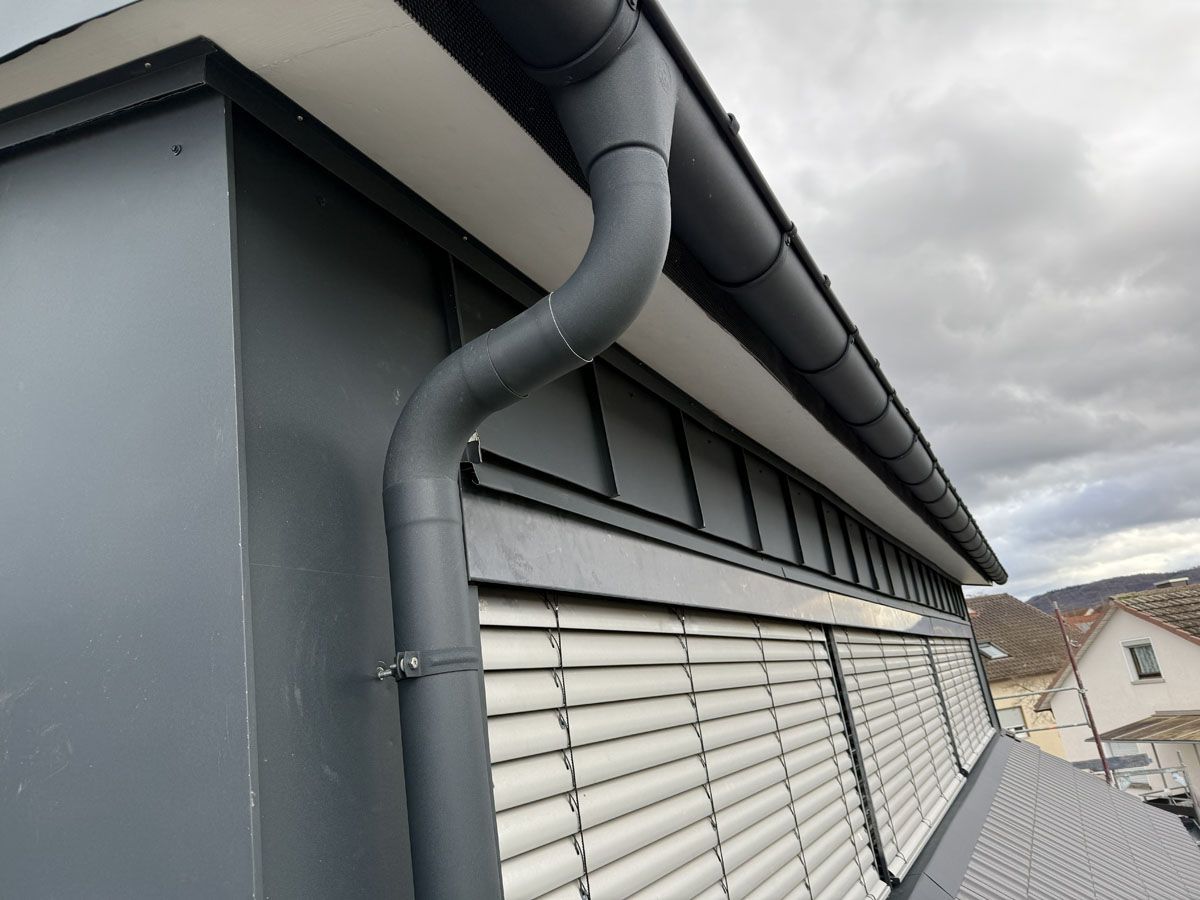 Ein graues Fallrohr an der Seite eines modernen Gebäudes mit grauer Fassadenverkleidung und außenliegenden Jalousien.