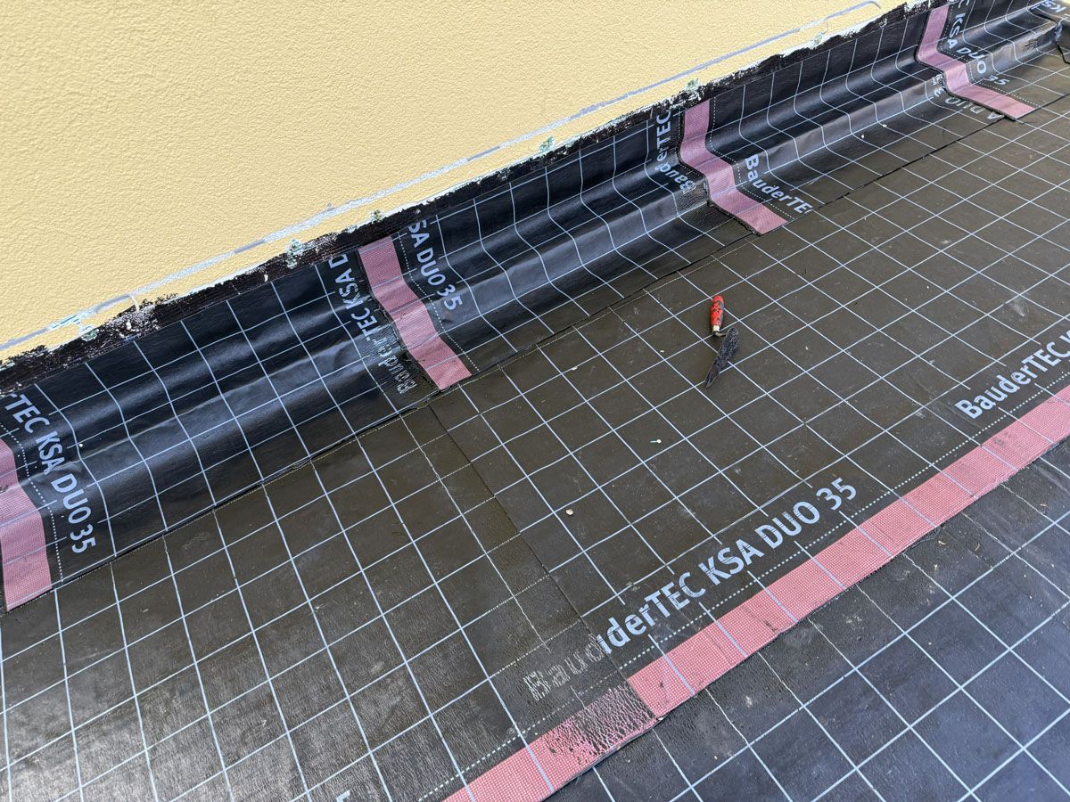 Schwarze, wasserdichte Dachmembran mit Gittermarkierungen und rosa Klebestreifen, die entlang einer strukturierten gelben Wand angebracht wurde.