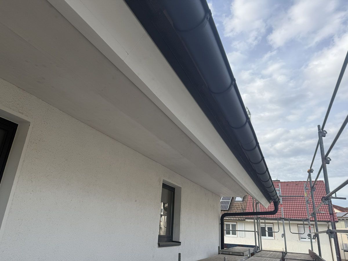 Ansicht aus der Froschperspektive auf die Fassade eines weißen Stuckhauses mit tiefem Dachüberstand, schwarzen Dachrinnen und modernen Fenstern.